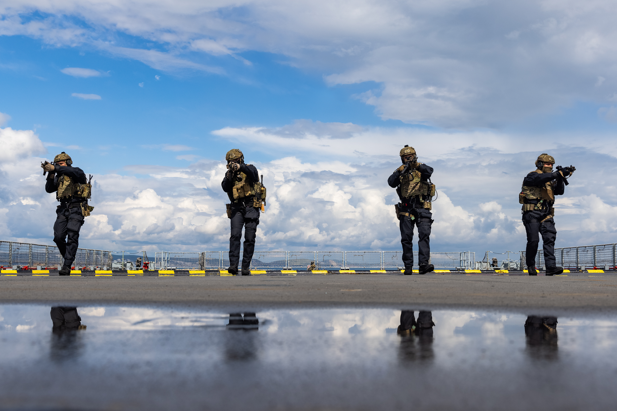 Four Royal Marines in camouflage uniforms with rifles standing on a deck against a cloudy sky.